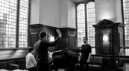 A black and white image of a singe and pianist being coached by a third persosn who has their arm in the air